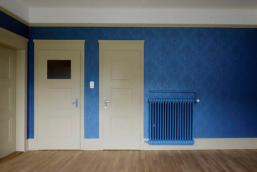 Wohnraum mit kräftig blauer Mustertapete, zwei frisch lackierten Kassettentüren in Cremeweiss und blau lackiertem Heizkörper