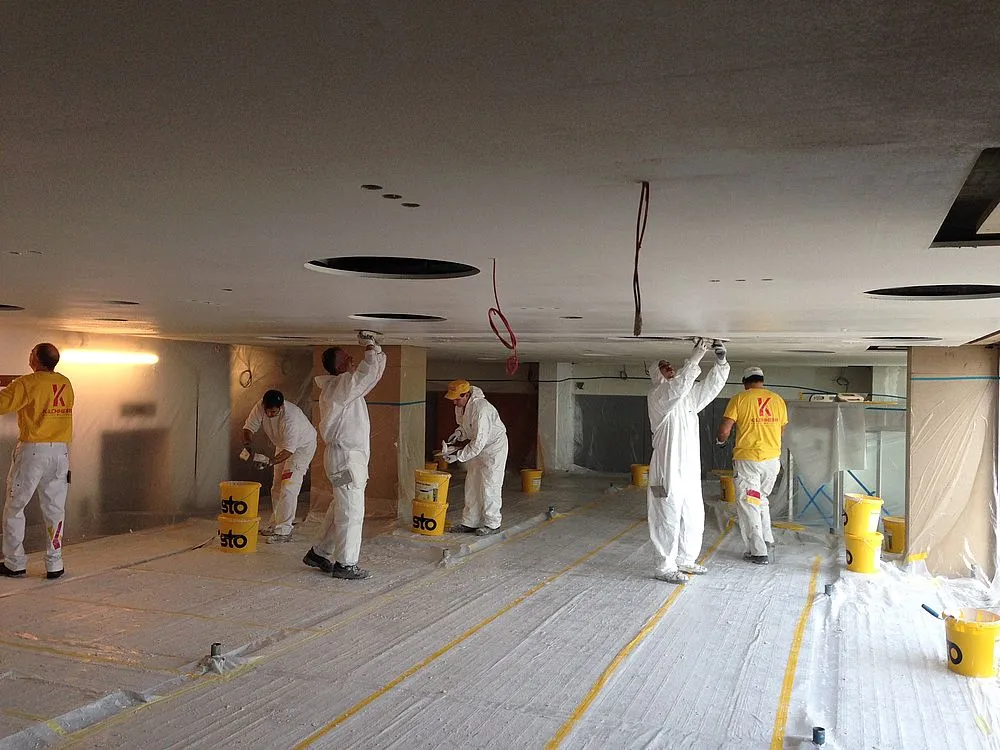 Kilchherr-Team in gelben Shirts und weissen Overalls beim Verputzen einer Decke, auf dem abgedeckten Boden stehen gelbe Farbkübel